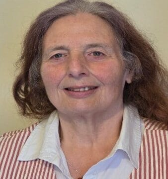 Margaret Graham, a board member, is an older woman with shoulder-length brown hair, wearing a white collared shirt and a red-and-white striped vest, smiling at the camera.