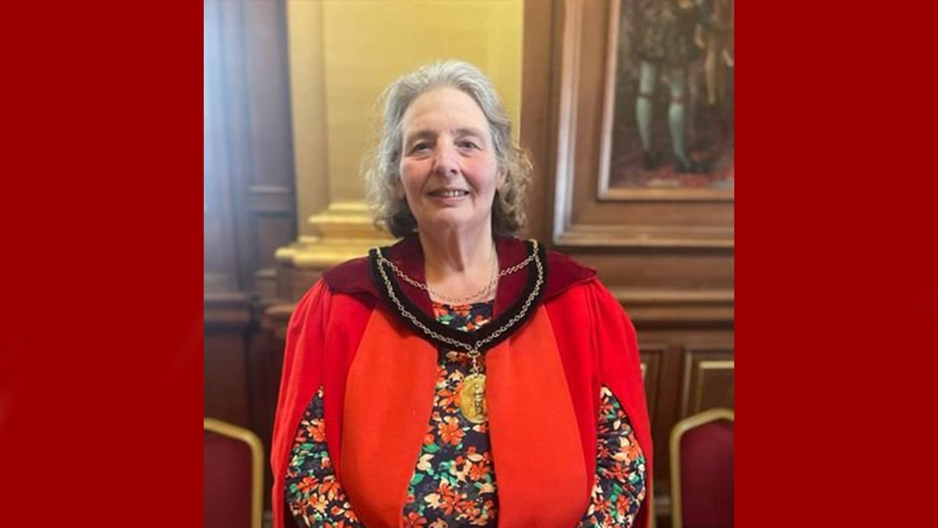 Margaret Graham, a member of the Capital Theatres Board, stands indoors before wooden paneling and framed artwork, dressed in a ceremonial red robe with a chain and medallion.