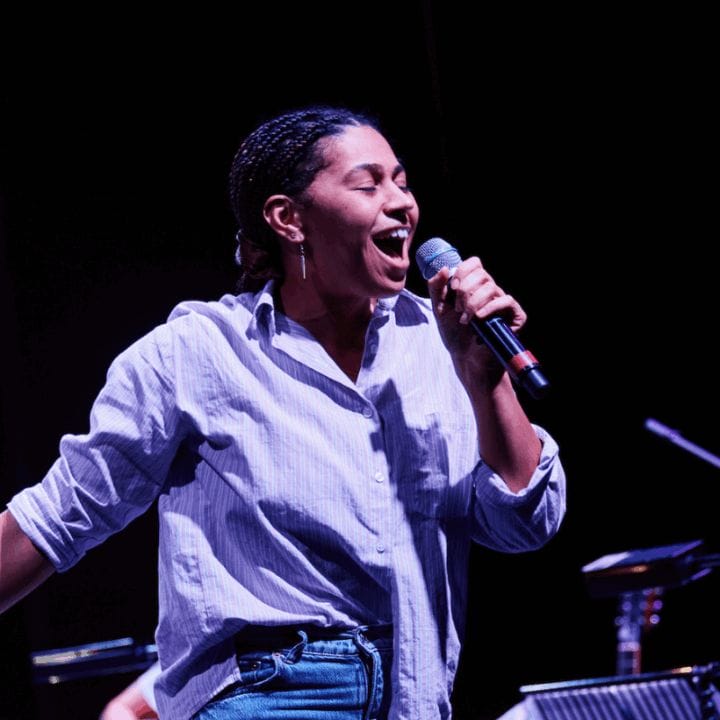 A person in a light shirt and jeans holds a microphone and sings or speaks on stage, with music stands and a seated musician visible in the background.