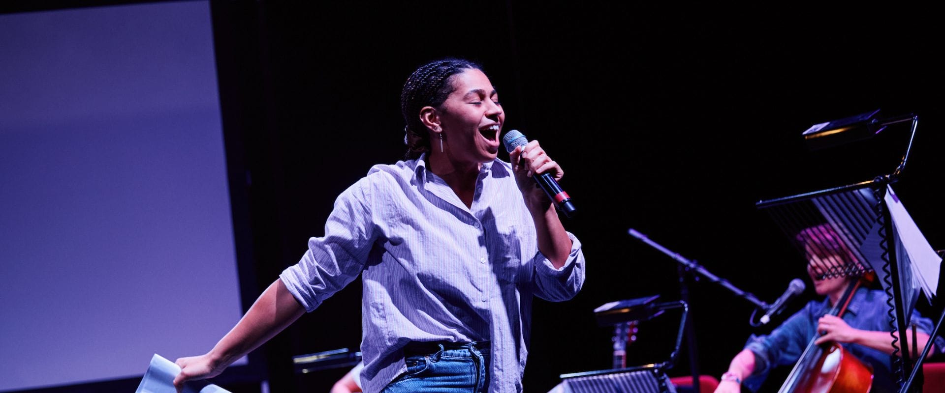 A person in a light shirt and jeans holds a microphone and sings or speaks on stage, with music stands and a seated musician visible in the background.