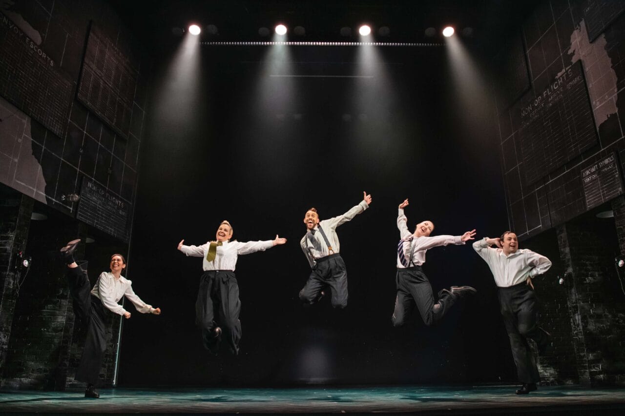Five people in dress shirts and suspenders are mid-jump, performing a synchronized dance routine on a stage under bright stage lights.