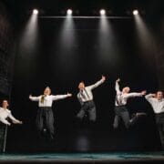 Five people in dress shirts and suspenders are mid-jump, performing a synchronized dance routine on a stage under bright stage lights.