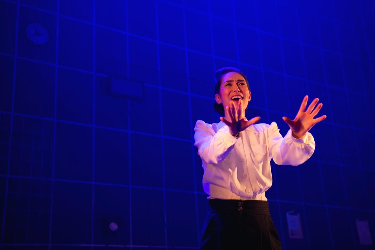 A person in a white shirt and dark pants stands on stage under blue lighting, gesturing with both hands and appearing to speak or sing with emotion.