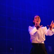 A person in a white shirt and dark pants stands on stage under blue lighting, gesturing with both hands and appearing to speak or sing with emotion.