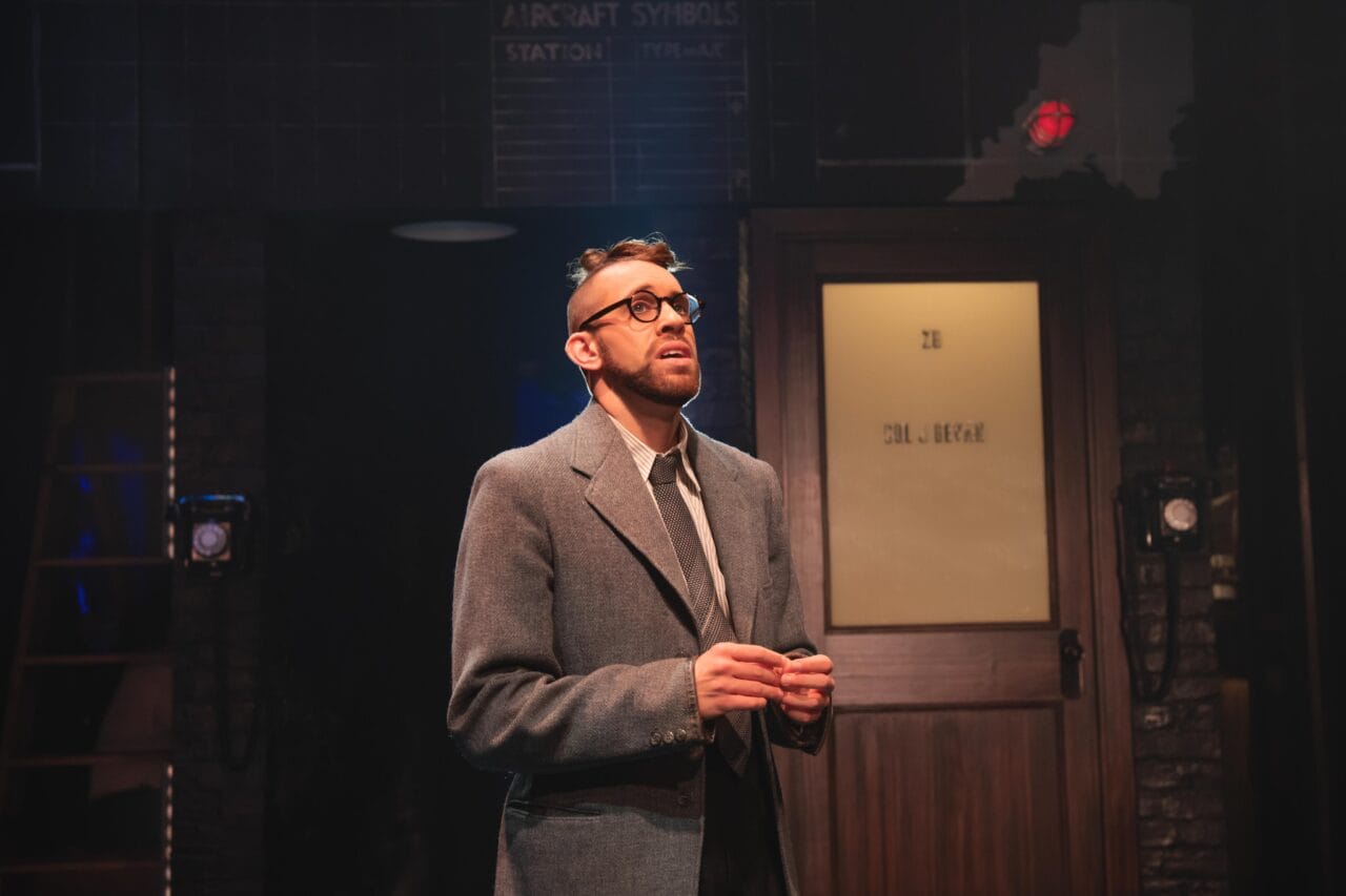 A man in a suit and glasses stands under stage lighting in front of a door labeled "28" with a serious expression, appearing to perform in a theatrical setting.