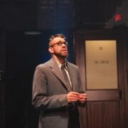 A man in a suit and glasses stands under stage lighting in front of a door labeled "28" with a serious expression, appearing to perform in a theatrical setting.