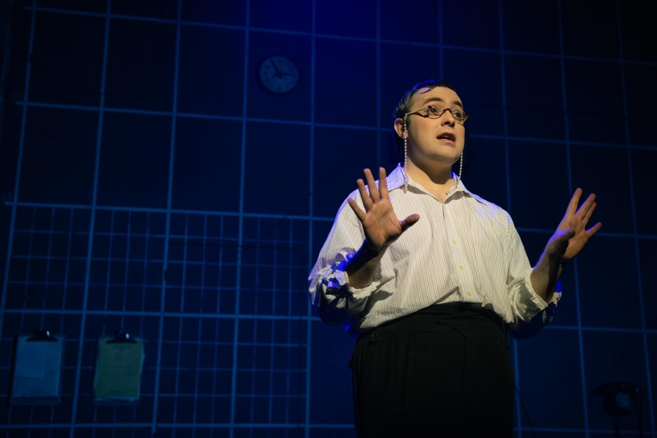 Person wearing glasses and a white shirt stands on stage with hands raised, in front of a blue grid-patterned wall with a clock and two clipboards visible.