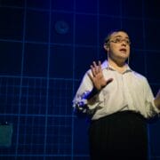 Person wearing glasses and a white shirt stands on stage with hands raised, in front of a blue grid-patterned wall with a clock and two clipboards visible.