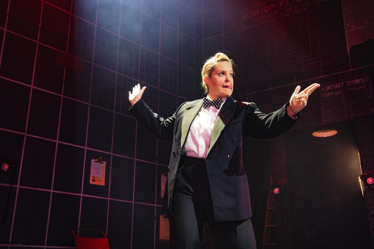 A person in a suit and bow tie stands on stage under dramatic lighting, arms outstretched, with a background of grid lines and red light.