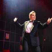 A person in a suit and bow tie stands on stage under dramatic lighting, arms outstretched, with a background of grid lines and red light.