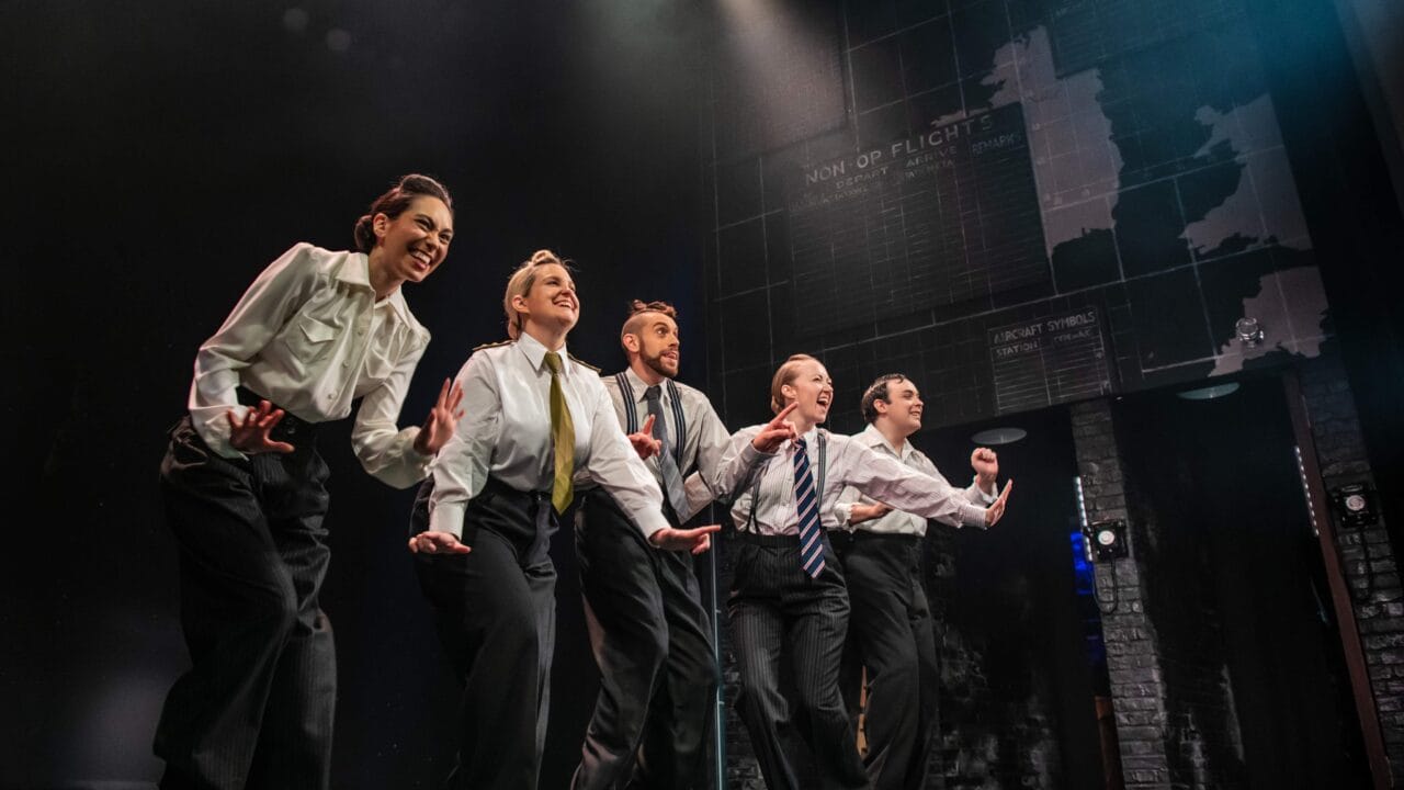 Five actors in suits perform energetically on a dimly lit stage, with a backdrop featuring maps and text.