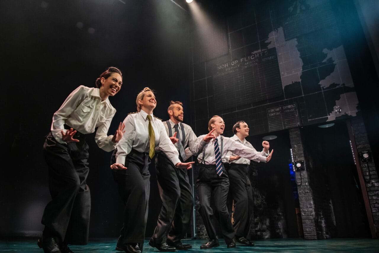 Five actors in suits perform energetically on a dimly lit stage, with a backdrop featuring maps and text.