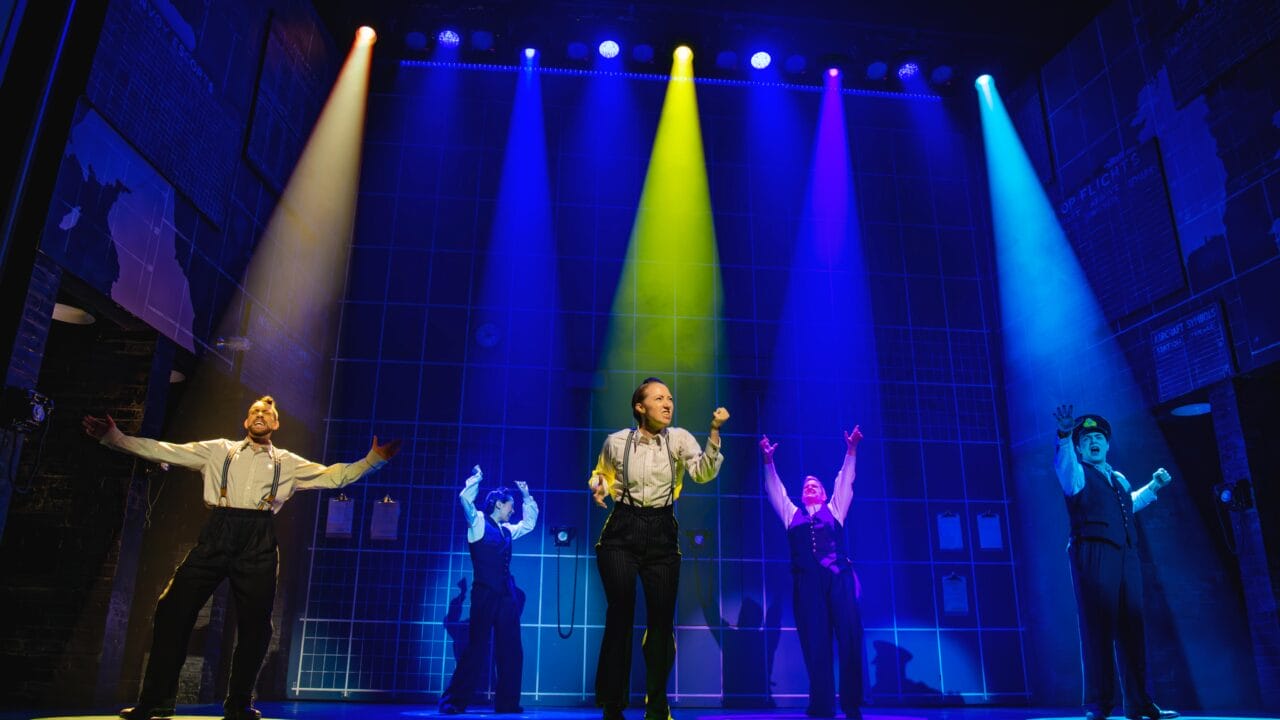 Four performers stand on stage under colorful spotlights, each with arms raised, against a backdrop featuring a grid pattern and brick walls.