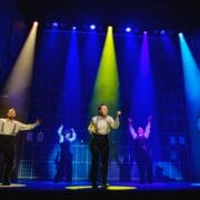 Four performers stand on stage under colorful spotlights, each with arms raised, against a backdrop featuring a grid pattern and brick walls.