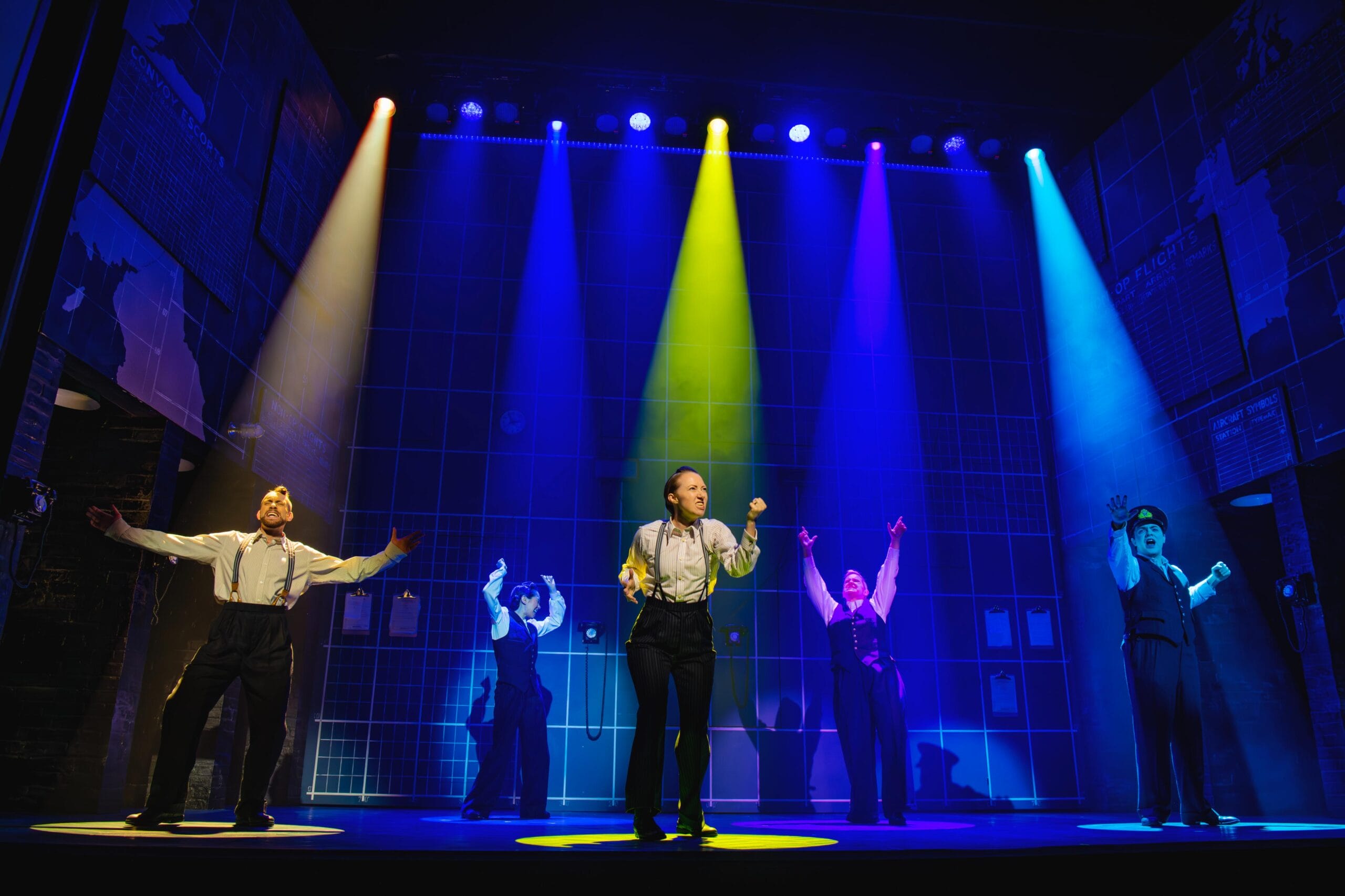 Four performers stand on stage under colorful spotlights, each with arms raised, against a backdrop featuring a grid pattern and brick walls.