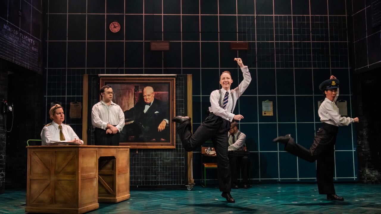 Five actors in business attire perform energetically on a stage set resembling an office, with a large portrait on the back wall and a person seated at a desk.