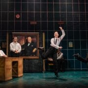 Five actors in business attire perform energetically on a stage set resembling an office, with a large portrait on the back wall and a person seated at a desk.