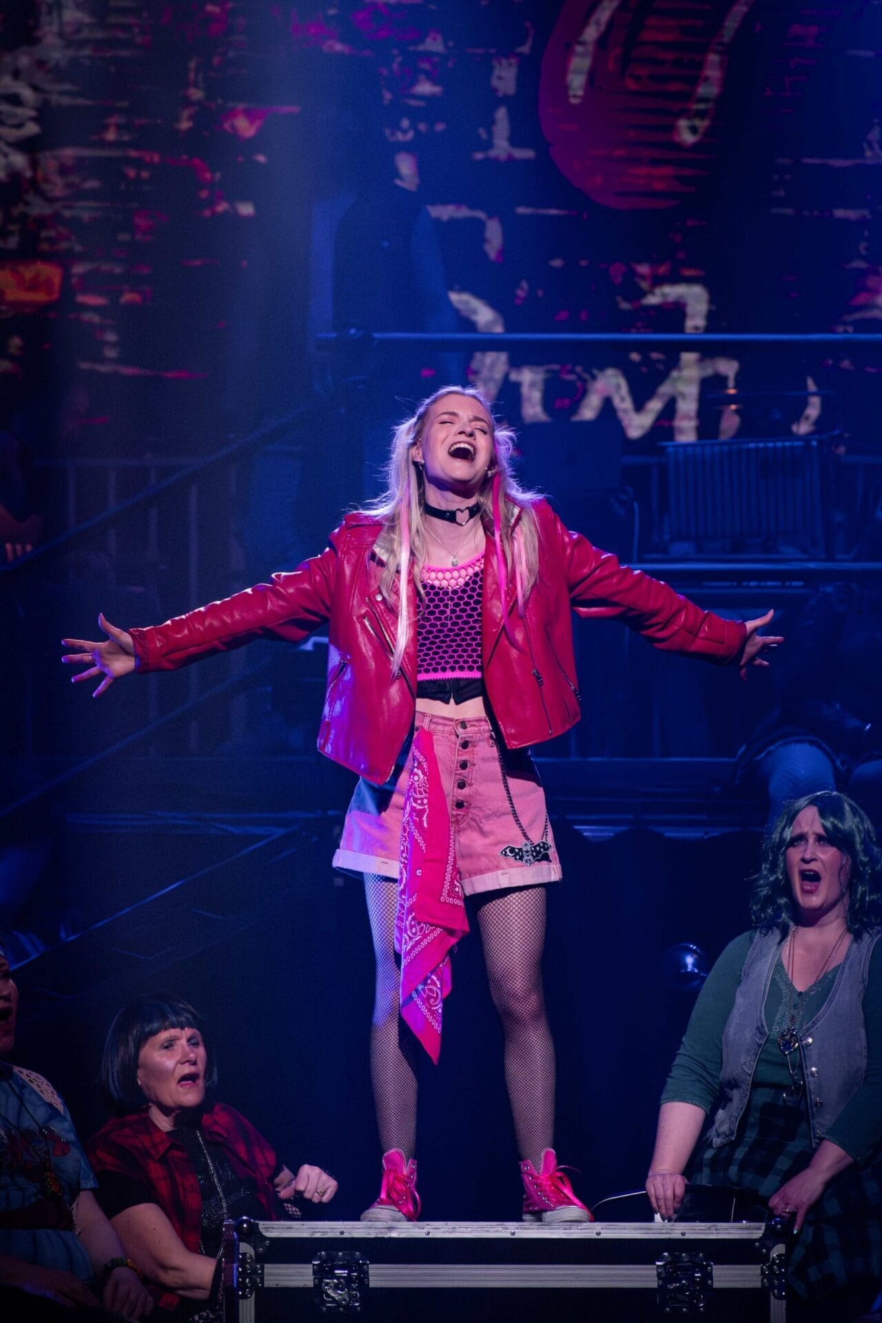 A young woman in a pink skirt, pink jacket, and pink shoes stands on a platform with arms outstretched, singing passionately to "We Will Rock You" on stage with other performers seated below her under dramatic stage lighting.