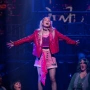 A young woman in a pink skirt, pink jacket, and pink shoes stands on a platform with arms outstretched, singing passionately to "We Will Rock You" on stage with other performers seated below her under dramatic stage lighting.