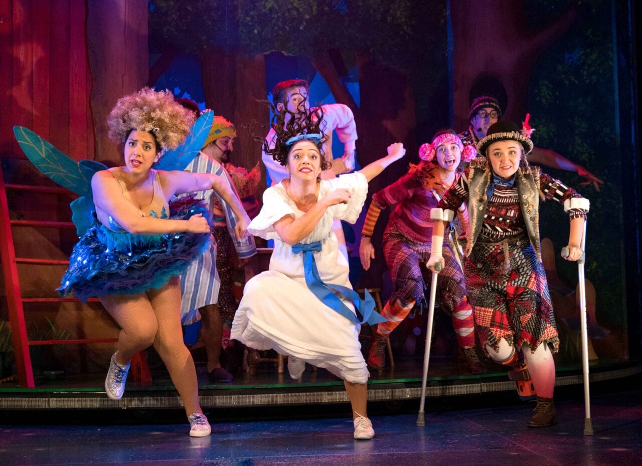 A group of actors in colourful costumes perform energetically on stage, with a forest backdrop and theatrical lighting.