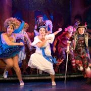 A group of actors in colourful costumes perform energetically on stage, with a forest backdrop and theatrical lighting.
