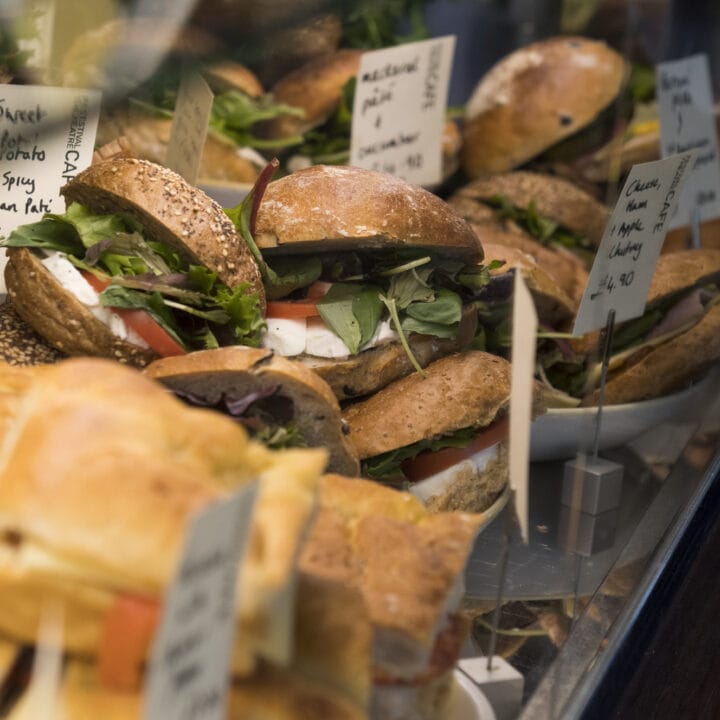 Display of assorted sandwiches and filled rolls behind a glass counter, with handwritten signs listing names and prices.