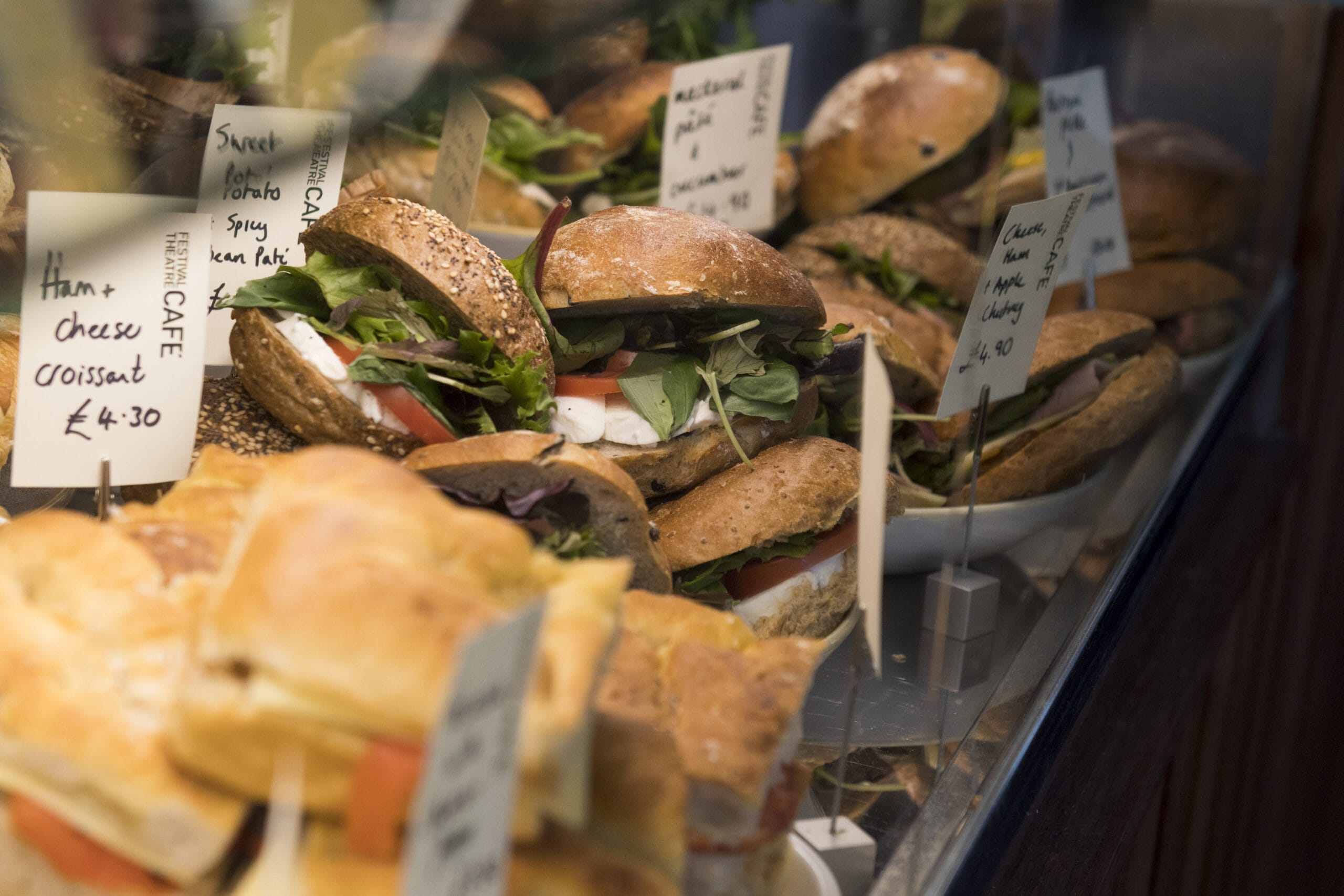 Display of assorted sandwiches and filled rolls behind a glass counter, with handwritten signs listing names and prices.