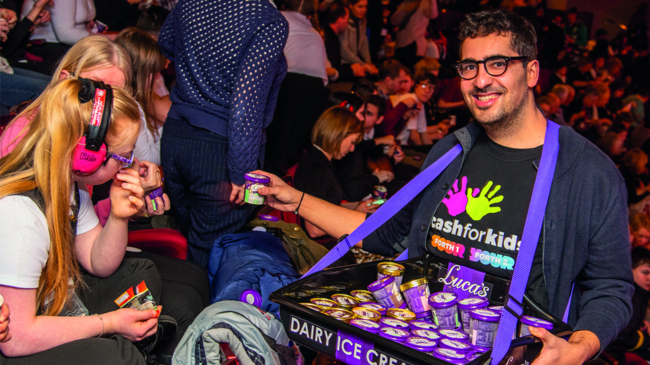 A man distributes small tubs of dairy ice cream from a tray to people seated in a crowded audience; one person wears noise-cancelling headphones.