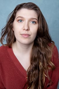 A woman with long brown hair and blue eyes wearing a red sweater, looking directly at the camera against a blue background.
