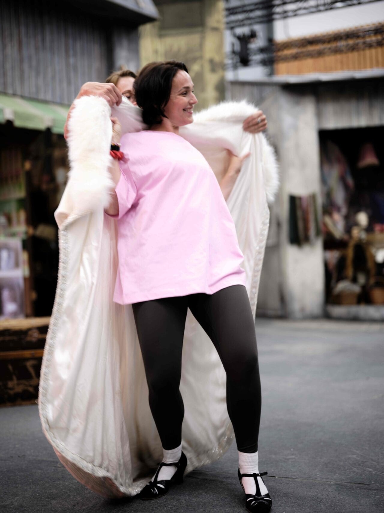A woman in a pink shirt, black leggings, and dance shoes is being helped into a long white fur-trimmed coat in an urban outdoor setting.