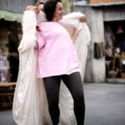 A woman in a pink shirt, black leggings, and dance shoes is being helped into a long white fur-trimmed coat in an urban outdoor setting.