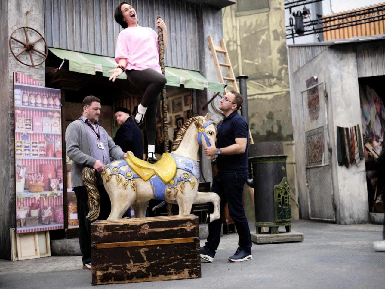 A woman enthusiastically poses on a carousel horse prop, while three men stand nearby in a film or set environment.