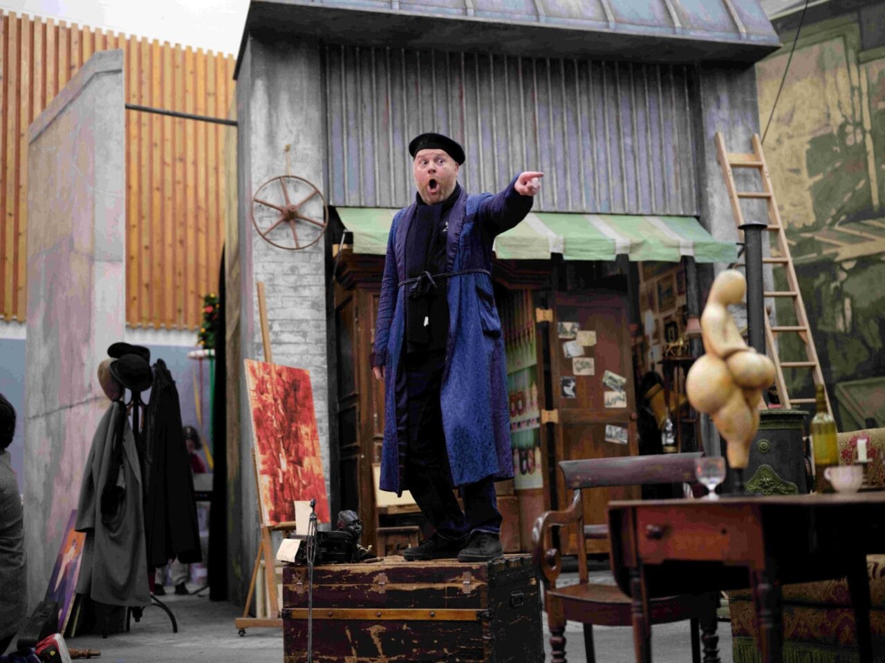 A man in a blue coat and black beret stands on a wooden crate, pointing and shouting, in front of a set resembling an old shop with various props and furniture.