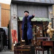 A man in a blue coat and black beret stands on a wooden crate, pointing and shouting, in front of a set resembling an old shop with various props and furniture.