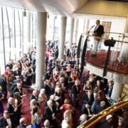 A large crowd gathers in a spacious, modern venue as a man speaks at a podium on an upper level. The event appears formal, with attendees dressed in suits and dresses.