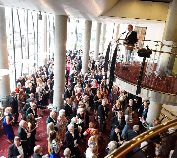 A large crowd gathers in a spacious, modern venue as a man speaks at a podium on an upper level. The event appears formal, with attendees dressed in suits and dresses.