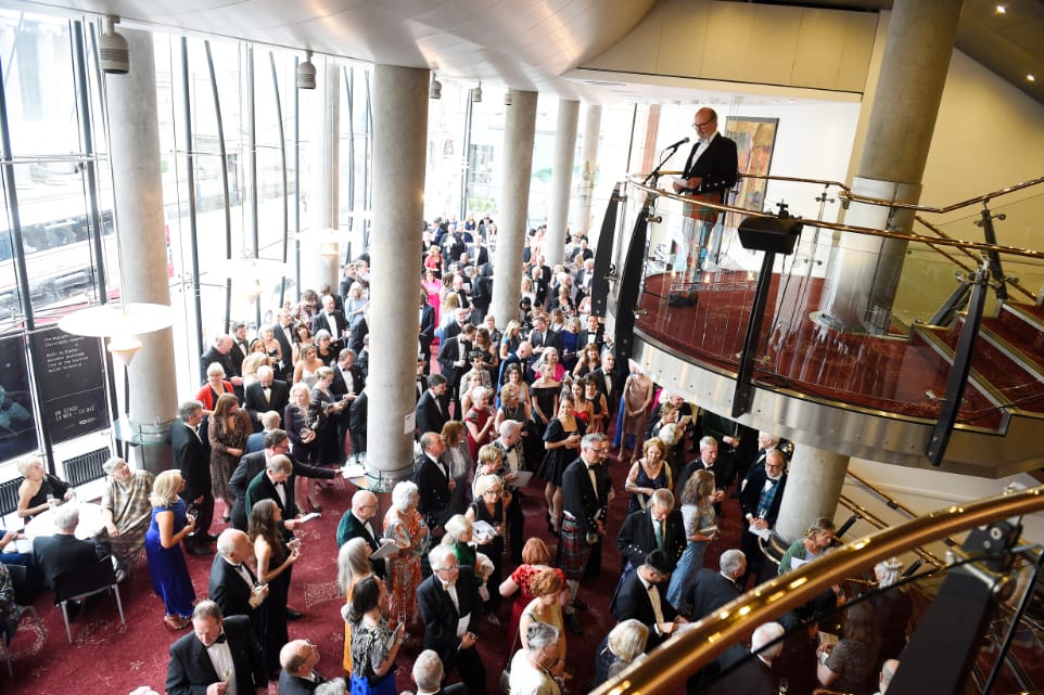 A large crowd gathers in a spacious, modern venue as a man speaks at a podium on an upper level. The event appears formal, with attendees dressed in suits and dresses.