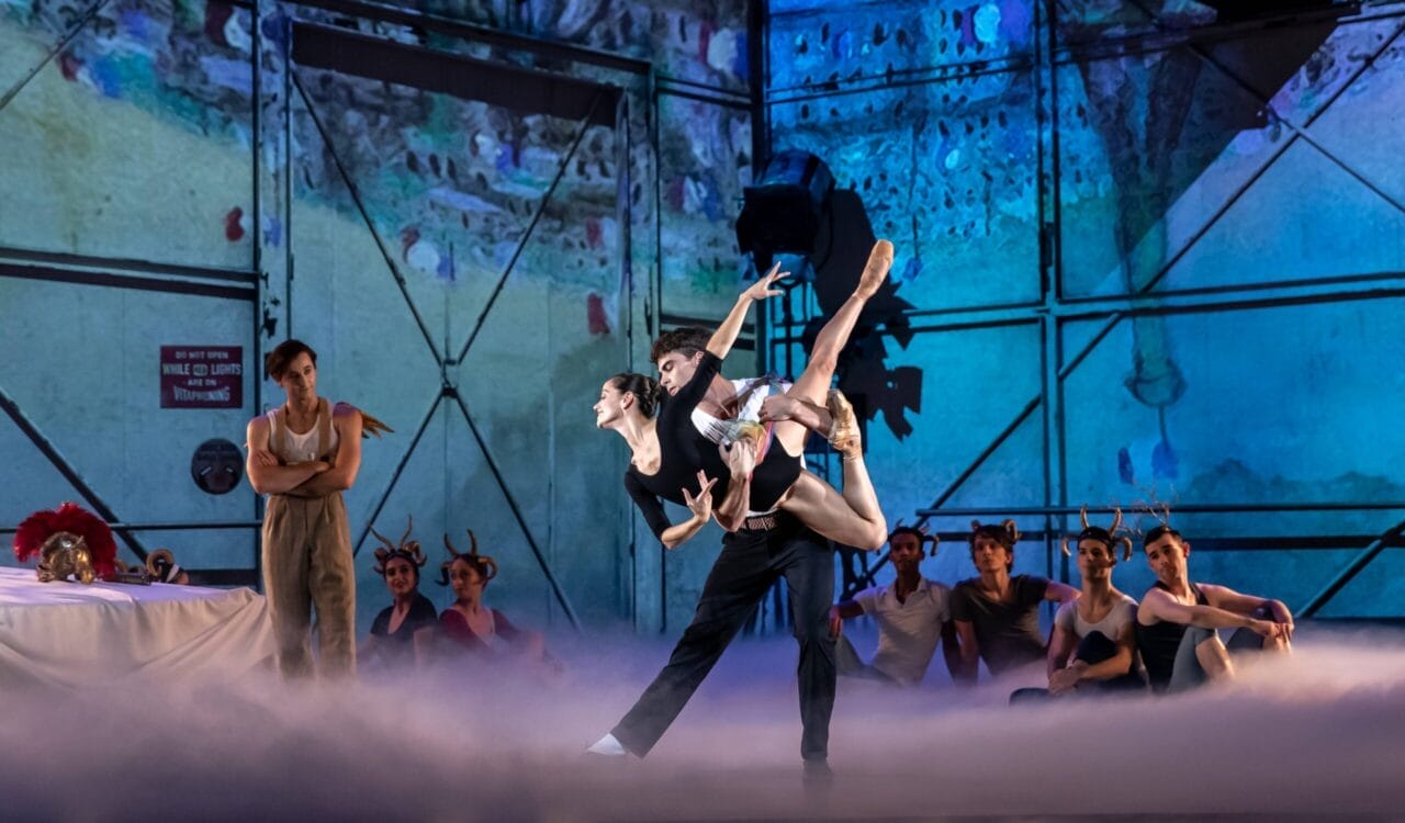 Two ballet dancers perform a lift center stage, surrounded by watching performers and stage fog, with a colorful backdrop and industrial set design.
