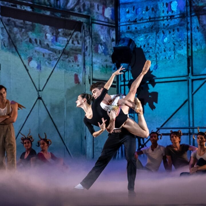 Two ballet dancers perform a lift center stage, surrounded by watching performers and stage fog, with a colorful backdrop and industrial set design.