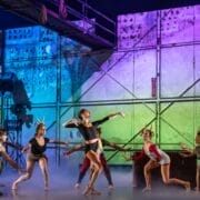 A group of dancers in costumes rehearse on a stage with colorful, industrial-style scenery and dramatic lighting.