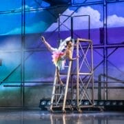 A ballerina in a colorful costume performs an arabesque pose on a metal scaffolding under theatrical stage lighting and backdrop.