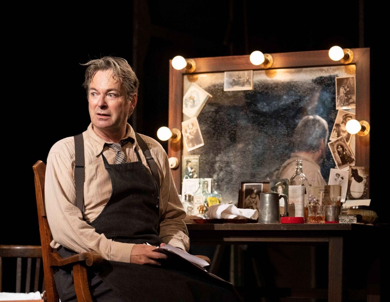 A man in an apron sits in front of a cluttered, lit vanity mirror with photos and bottles, holding papers and looking to the side.