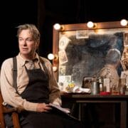 A man in an apron sits in front of a cluttered, lit vanity mirror with photos and bottles, holding papers and looking to the side.