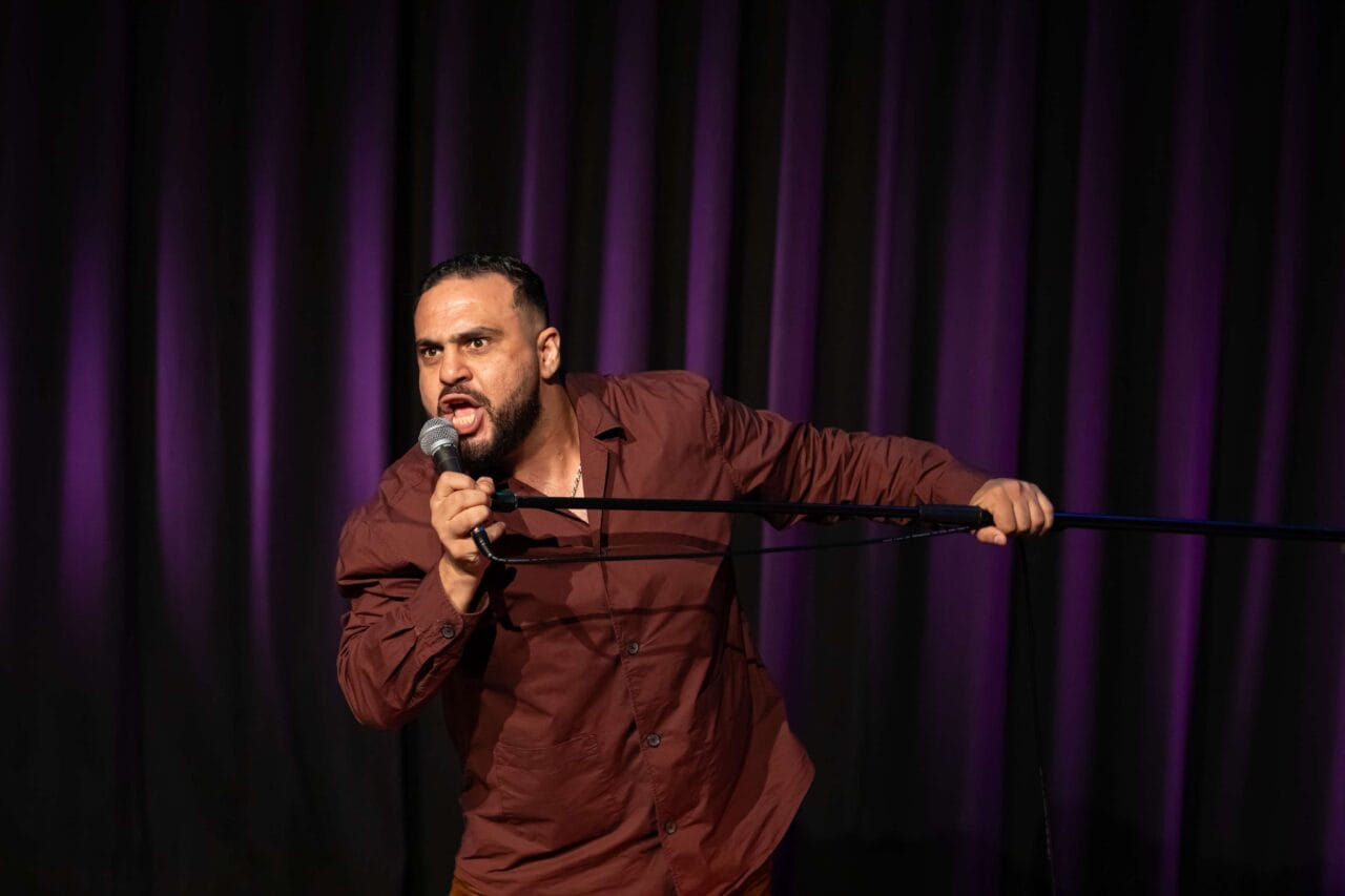 A man in a brown shirt passionately speaks into a microphone on stage, holding a microphone stand, with dark purple curtains in the background.