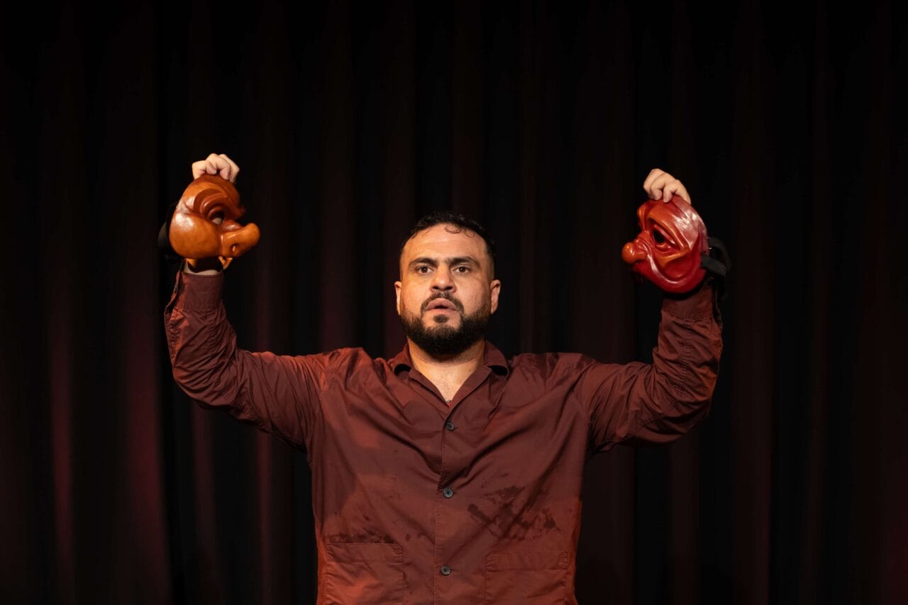 A man in a maroon shirt stands in front of a dark curtain, holding two theatrical masks, one in each hand, raised at shoulder height.