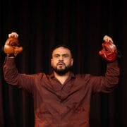 A man in a maroon shirt stands in front of a dark curtain, holding two theatrical masks, one in each hand, raised at shoulder height.