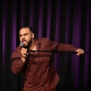 A man in a brown shirt passionately speaks into a microphone on stage, holding a microphone stand, with dark purple curtains in the background.