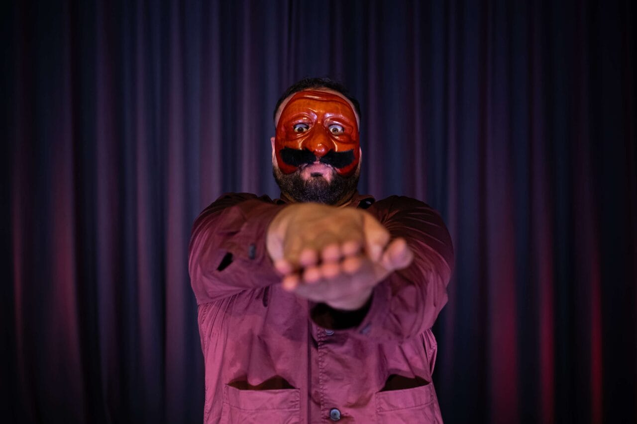 A person wearing a red theatrical mask and maroon shirt stands in front of dark curtains, holding their arms straight out with palms together, facing the camera.