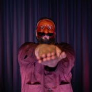 A person wearing a red theatrical mask and maroon shirt stands in front of dark curtains, holding their arms straight out with palms together, facing the camera.