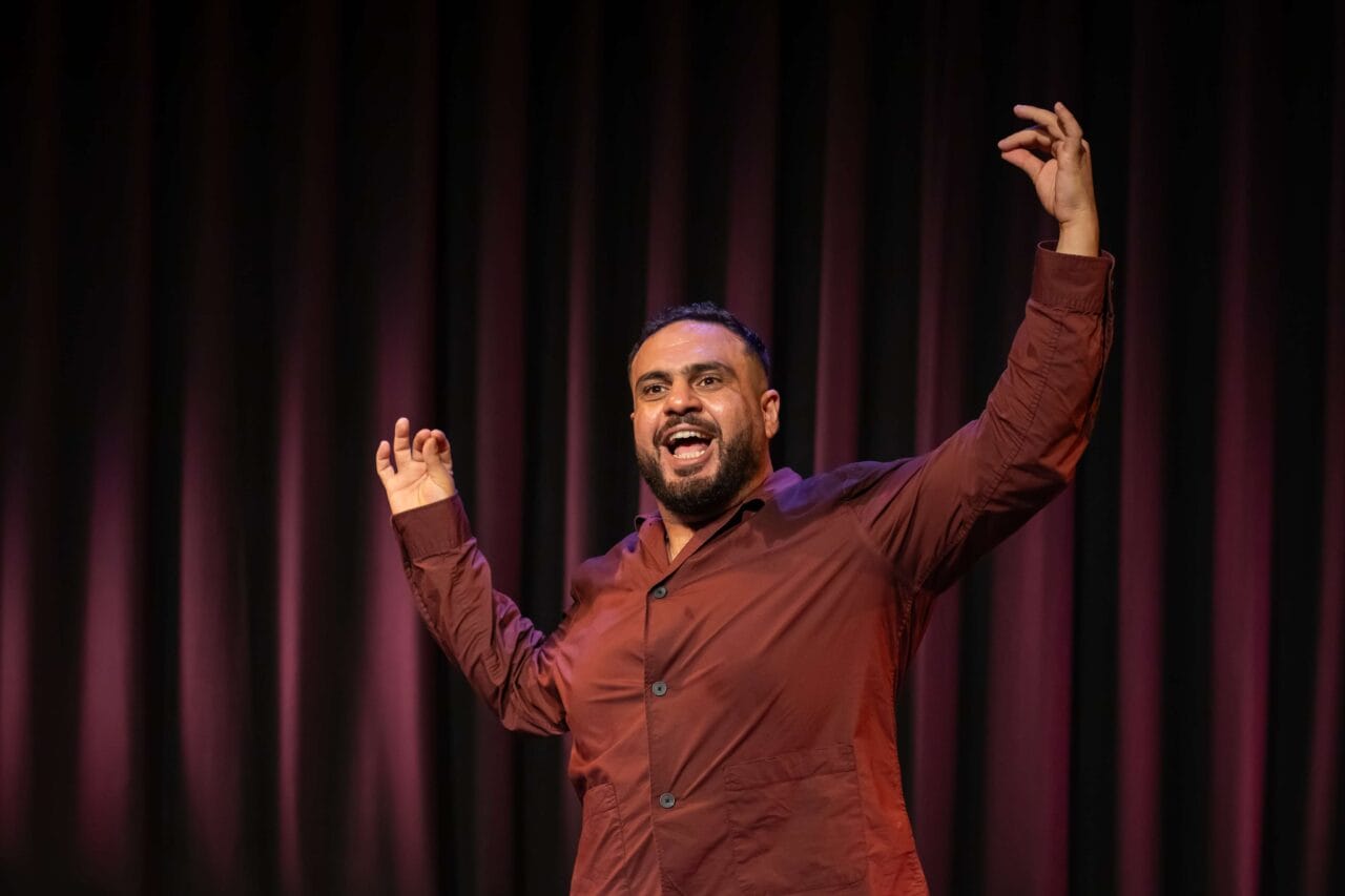 A man in a maroon shirt gestures with raised arms and open hands while speaking or performing on a stage with a dark curtain backdrop.